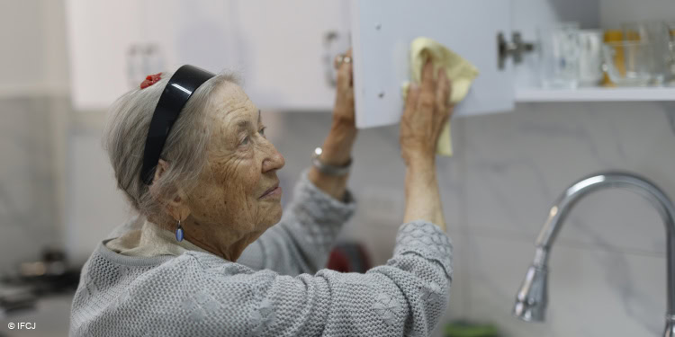 Elderly woman cleaning a kitchen cabinet, representing senior care and community support initiatives by IFCJ.