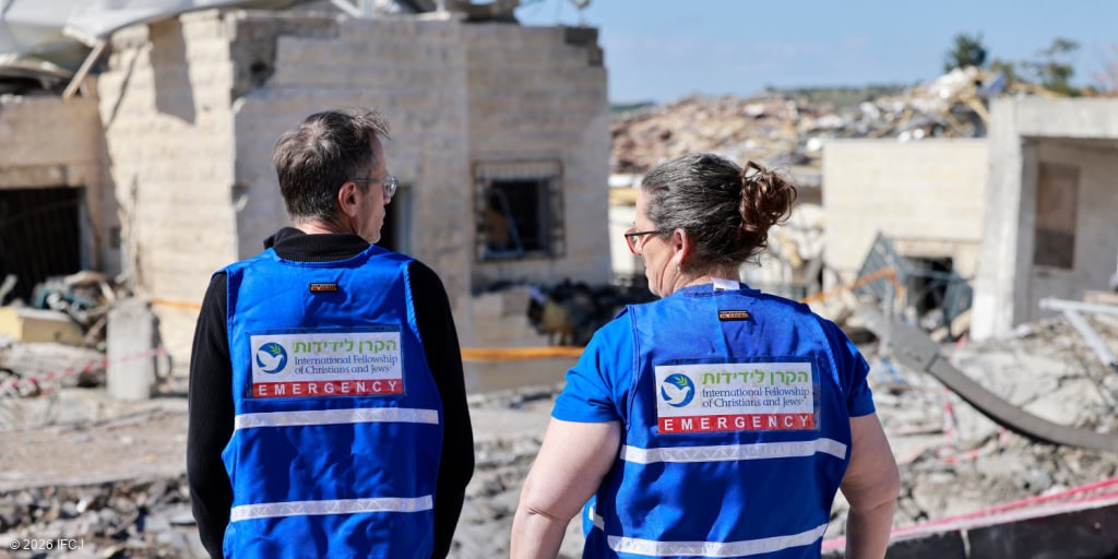Two emergency relief workers in blue uniforms providing aid in a disaster-stricken area with damaged buildings and debris.