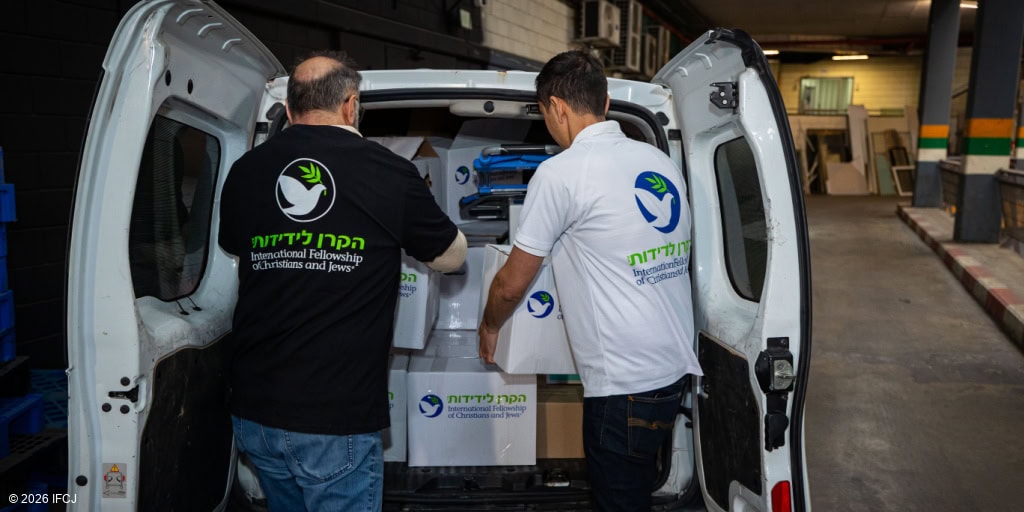 Van seen from rear with back doors open, white branded logo food boxes inside - man in black branded logo t-shirt and glasses holding a food box, Gadi Dan (IFCJ Staff) in white branded logo shirt, holding food box - loading boxes into the van