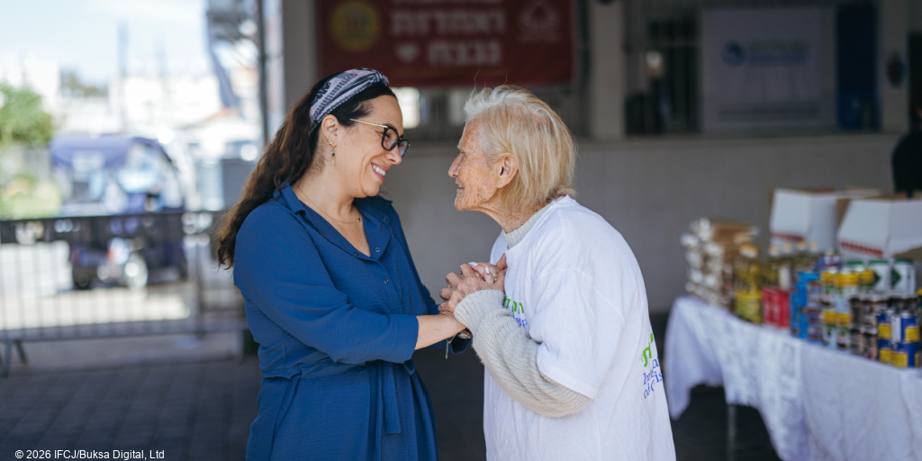 Yael Eckstein, blue dress, glasses - standing outside - talking to elderly woman, blonde hair, volunteer in branded logo t-shirt - holding hands, smiling