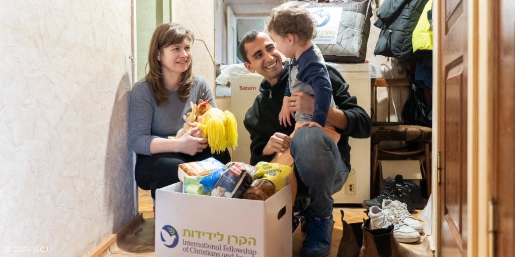 Gadi Dan (IFCJ Staff) and Yelena smiling at Carolina - black branded logo jacket - white branded logo food box - hallway - Yelena, dark gray shirt, holding stuffed doll with yellow yarn hair - Carolina, light gray shirt with navy blue sleeves, light orange pants - winter blanket in branded logo storage bag