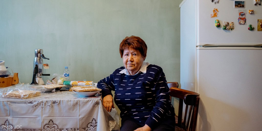 Elderly woman sitting at kitchen table - looking at camera - black and white sweater - table covered in white tablecloth with lace edge - matzah on table - white refrigerator