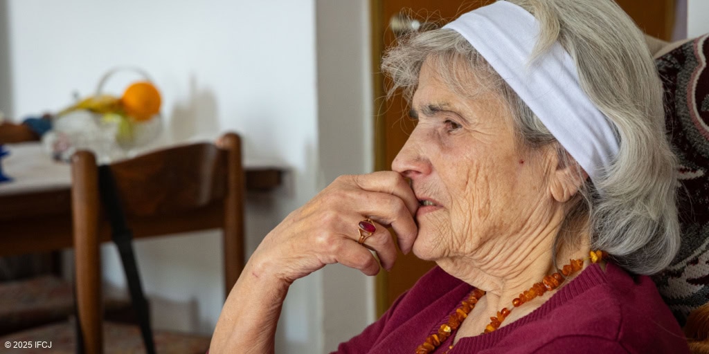 Elderly woman with gray hair, wearing a white headscarf and red sweater, sitting indoors with a contemplative expression, representing compassion and support for seniors.