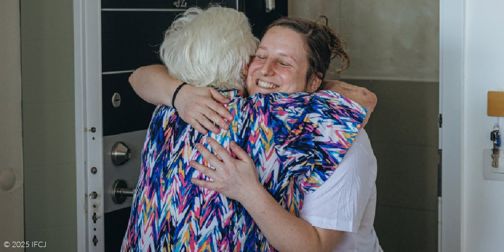 Elderly woman opening door, seen from behind - greeting IFCJ staff woman in open door - blue and pink patterned blouse, white T-shirt with IFCJ logo - smiling - hug