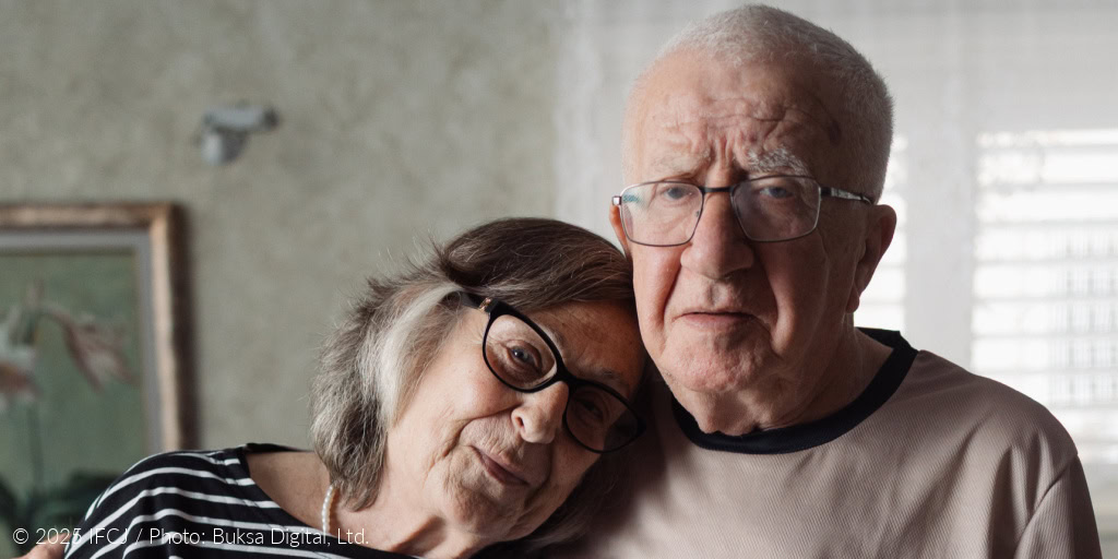Moshe Adler - elderly man, glasses, tan, gray shirt, standing - with his wife, Malka Adler - elderly woman in black and white striped shirt - arms around shoulder - gray couch - purple flowers - her head on his shoulder