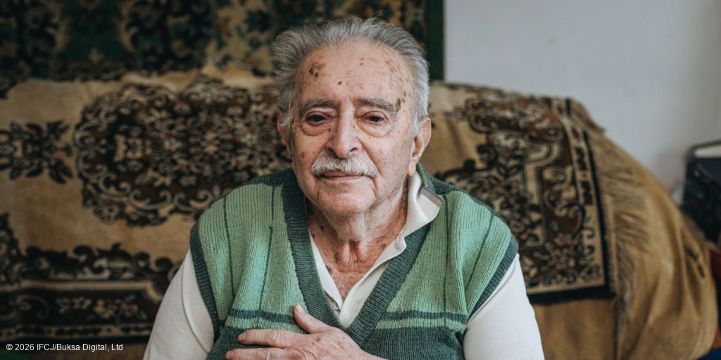 An elderly man with gray hair and a mustache sitting on a sofa, wearing a green sweater, in a cozy home setting, representing care and compassion.