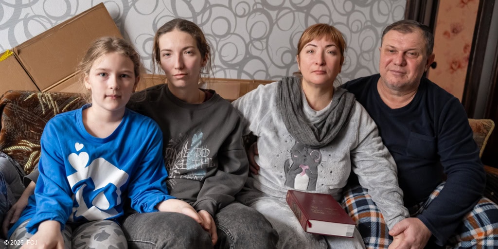 Family sitting on couch - 2 adult women, adult man in plaid pants, girl in blue and white shirt, gray swirls wallpaper, carboard boxes flattened - woman holding Torah book on her lap