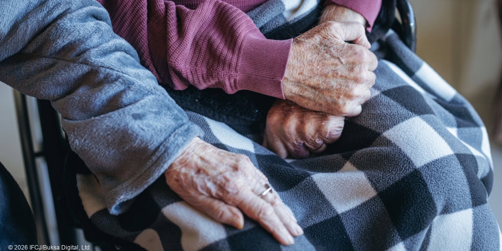 Dalia, woman in blue jacket - Pinhas, elderly man, purple long-sleeved t-shirt, black and white buffalo check blanket on his lap - her hand on his knee - no faces, focus on hands