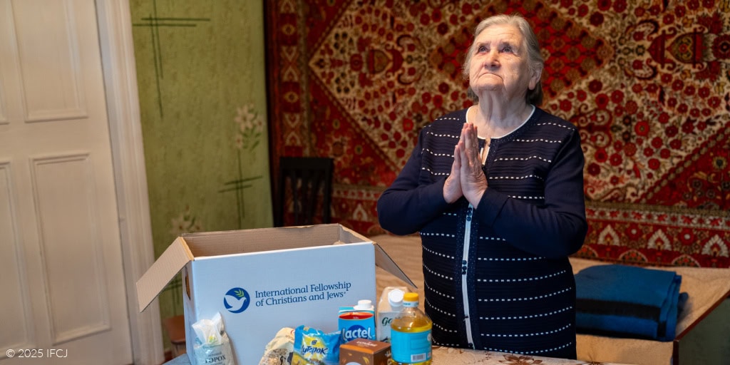 Elderly woman, black and white dress, looking up, holding hands in prayer - IFCJ logo food box on table with tan tablecloth - Oriental rug on wall - green wall - oranges, red pepper, milk, produce