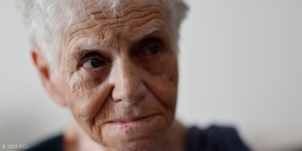 Close-up of elderly woman with short white hair, facing slightly to the side, with a softly furrowed brow, downturned mouth, and a steady gaze away from the camera, set against a softly lit, neutral indoor background.
