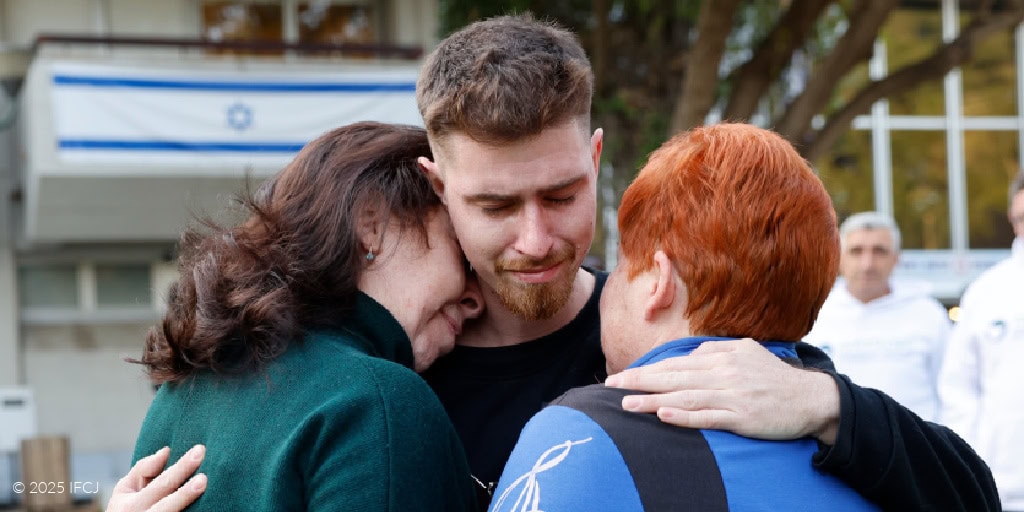 Vladimir Matveychuk and Svetlana Matveychuk - Tel Aviv, Israel - February 26, 2025 Injured Lone Soldier Visited by Ukrainian Mother and Aunt young adult man, black sweatsuit, outside, green grass, hugging adult woman with short red hair, light blue jacket, and adult woman with dark hair, dark green turtleneck dress, emotional - IFCJ staff standing in background, watching