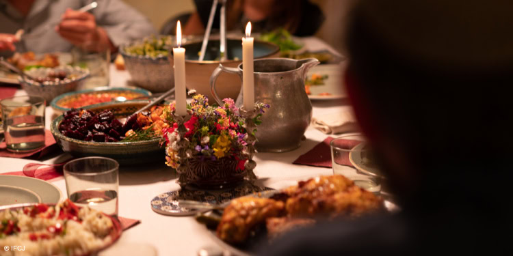 Stills from the model Shabbat production day, focus on the candles and flowers in the middle of the table, Liam and Sapir out of focus in the background, food on the table, rice, salad, plates, napkins, silverware, glass, pitcher