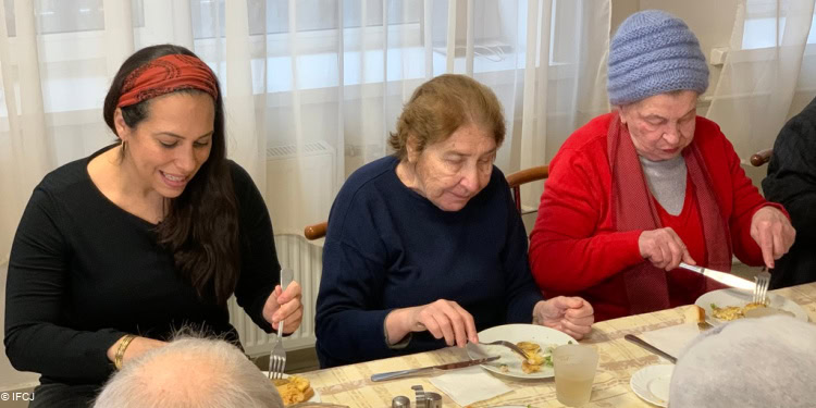 Dinner with holocaust survivors, DRTV Ukraine infomercial production January 14-24, 2020, Yael Eckstein sits at a dinner table with holocaust survivors, elderly women and elderly men, all looking at their plate of food, black shirt, orange shirt, blue hat, food, winter clothing