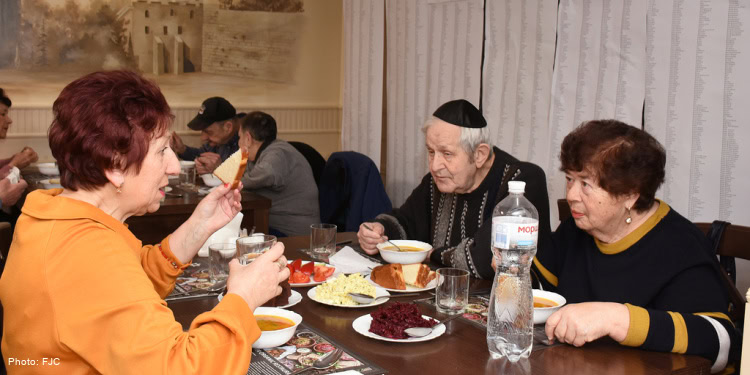 Patrons eating at a soup kitchen in Chernivtsi, Ukraine. Banner hanging on the wall with list of names of IFCJ donors who support the Jewish people. Banner reads "You are not alone. Enjoy this food, which is sent with love from a friend in America who cares for you." elderly men and women sitting at tables in soup kitchen, eating dinner