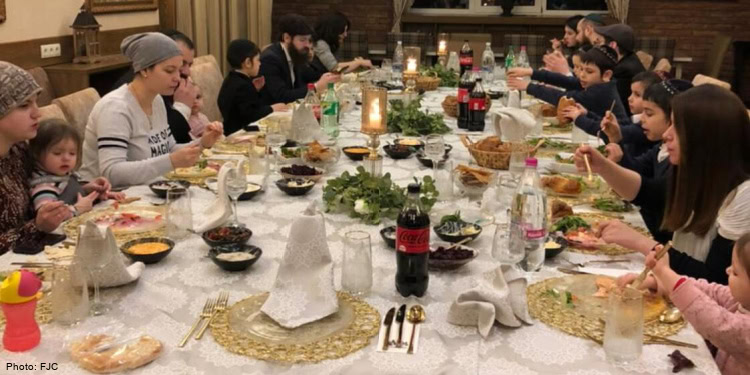 group of people at long dining room table eating dinner, white tablecloth, gold placemat, candles, headscarf, striped shirt, white shirt, black suit coat, hat, yarmulke, pink shirt