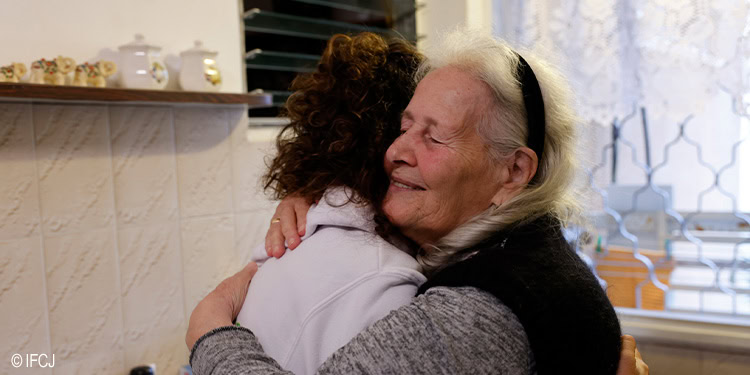 An elderly woman with white hair and a black headband hugging a young girl with curly hair in a warm, caring embrace indoors.
