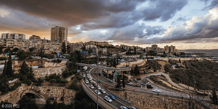 Sunset over Jerusalem with city buildings, roads, and traffic under dramatic clouds.