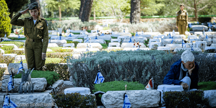 The image depicts a solemn scene at a cemetery during Yom Hazikaron, Israel Memorial Day. A young woman in military uniform salutes, while an elderly man sits by a grave, reflecting on the sacrifices of fallen soldiers and victims of terror. The cemetery is adorned with flags and memorial stones, emphasizing remembrance and national mourning.