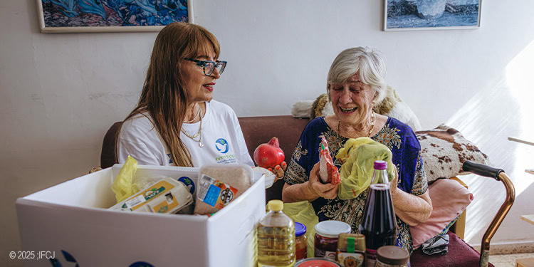 Elderly womand and IFCJ staff sitting on a sofa with a food box