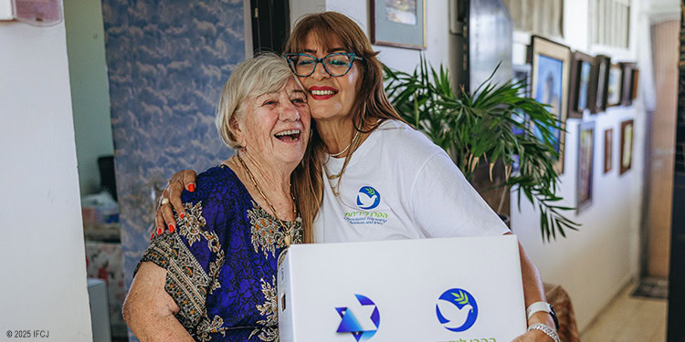 Woman wearing an IFCJ-branded shirt hugging an elderly woman with a food box