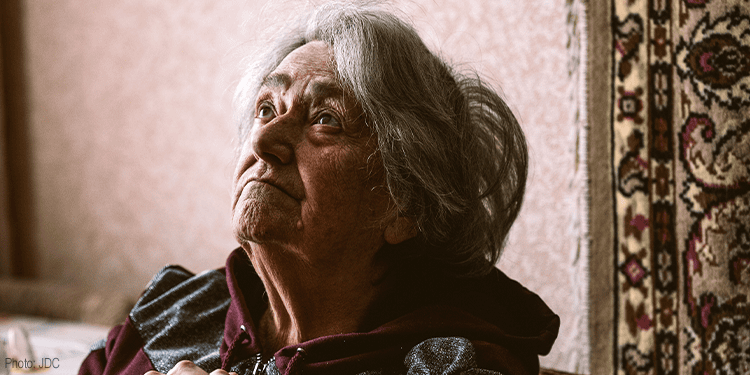 Close-up of an elderly woman with gray hair, looking contemplative and somber, sitting indoors against a textured wall and patterned carpet, illustrating humility and reflection.