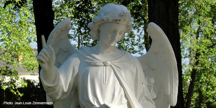 White angel sculpture with detailed wings and serene expression, set outdoors among green trees, symbolizing divine protection and spiritual guidance.