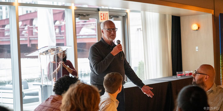 Man speaking into a microphone during a charity event at IFCJ headquarters, with attendees listening attentively in a well-lit room.