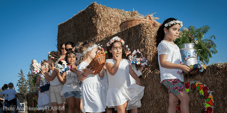 Children participating in a cultural festival, dressed in white dresses and floral crowns, holding flowers and traditional items, with hay bales and greenery in the background.