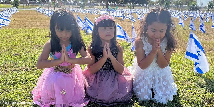 Three young girls in colorful dresses praying with hands together in a field decorated with Israeli flags, symbolizing hope and faith.