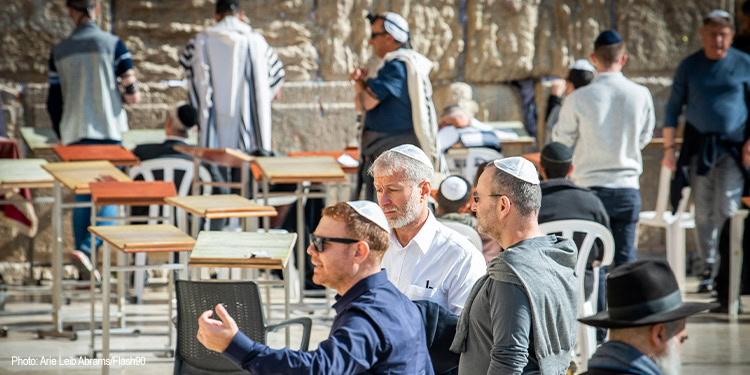 Jewish men praying at the Western Wall in Jerusalem, a sacred site for Jewish worship and pilgrimage, with visitors and tourists in the background.