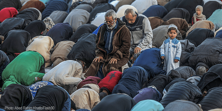 Muslim men and boys praying together during a religious gathering, with some women visible in the background, in an outdoor setting.
