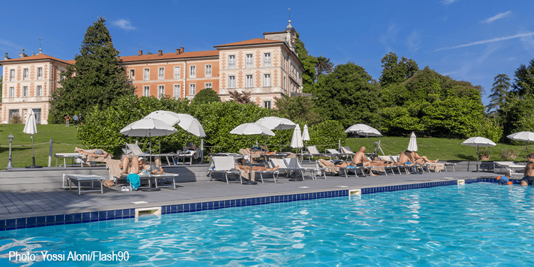 People relaxing by a luxurious outdoor hotel pool with umbrellas, surrounded by lush greenery and historic buildings under a clear blue sky.