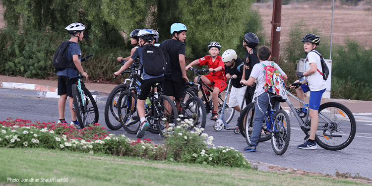 Group of children with helmets and backpacks gathered around their bikes on a paved path in a park setting.