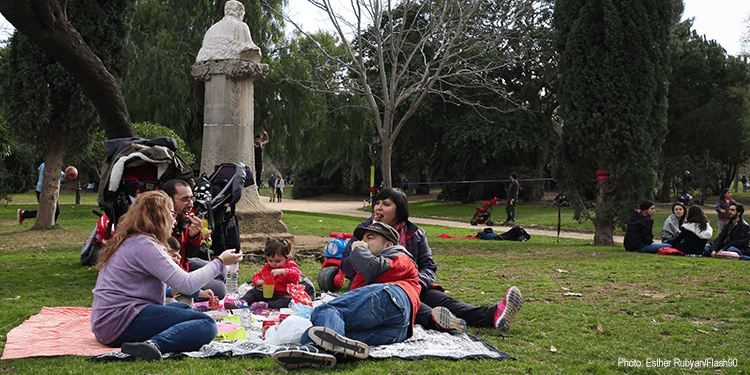 Families and children relaxing and playing in a park with trees and pathways, some sitting on blankets and others walking or talking.