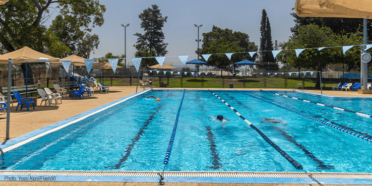 Outdoor public swimming pool with lane markers, umbrellas, and lounge chairs, surrounded by trees and clear sky, during daytime.