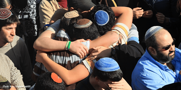 People embracing in a heartfelt mourning gathering, wearing traditional Jewish head coverings, expressing grief and solidarity during a memorial event.