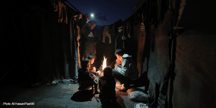 A family gathered around a small fire inside a dark tent, sharing a moment of warmth and connection during challenging times.