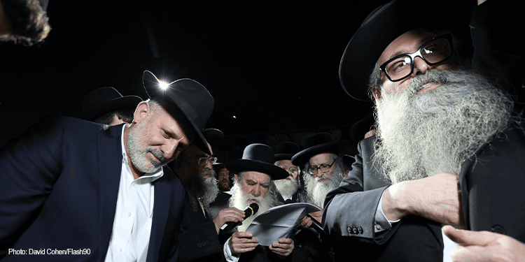 Group of Jewish men praying outdoors at night, wearing traditional black hats and suits, with focused expressions and prayer books in hand.