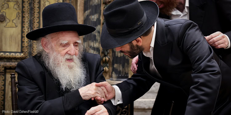 An elderly Jewish man with a long white beard and black hat shaking hands with a younger man in a black suit and hat, showing mutual respect.