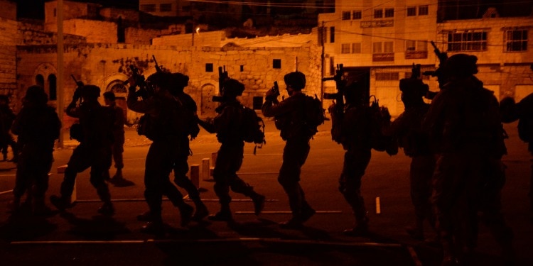 Several IDF soldiers in formation late at night with an orange light cast around them.