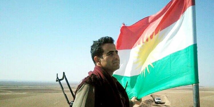 A man standing in the desert in front of a red, white, and green flag.