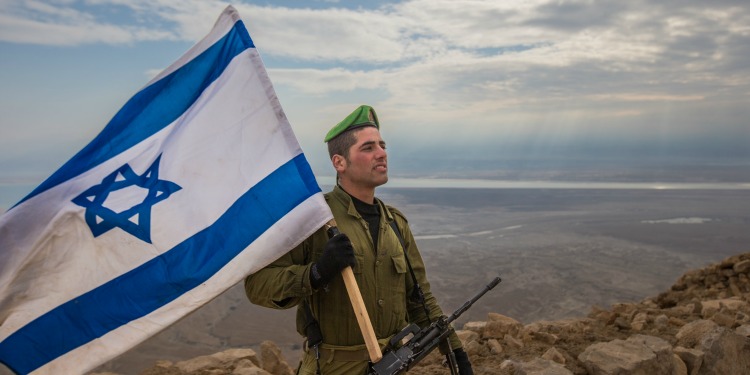 Soldier on top of a mountain while holding the Israeli flag.