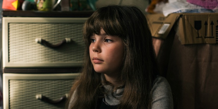 A young girl sitting on the floor looking off into the distance with several boxes behind her.