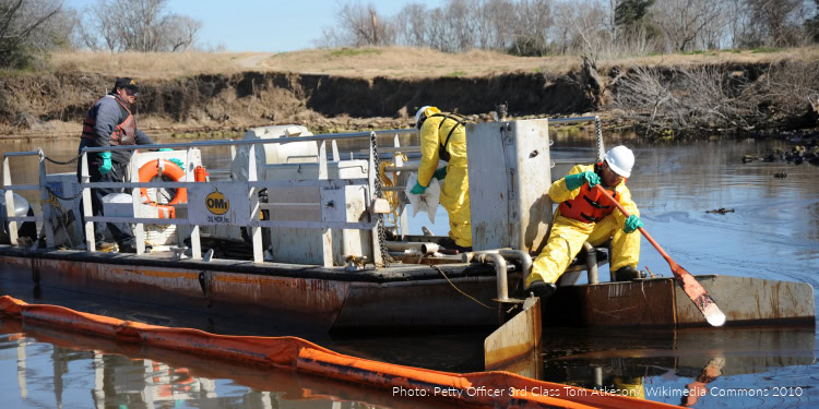Emergency responders in yellow protective suits clean an oil spill from a water body using specialized equipment and tools.