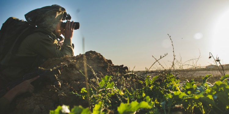 A soldier looking at the land ahead of him using binoculars.