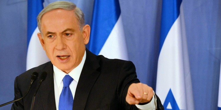 Bibi giving a speech at a podium with Israeli flags behind him.