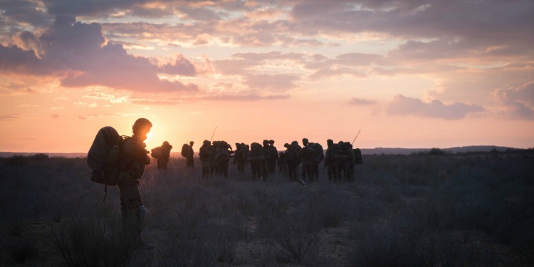 Soldiers walking in a field with a sunrise in front of them.