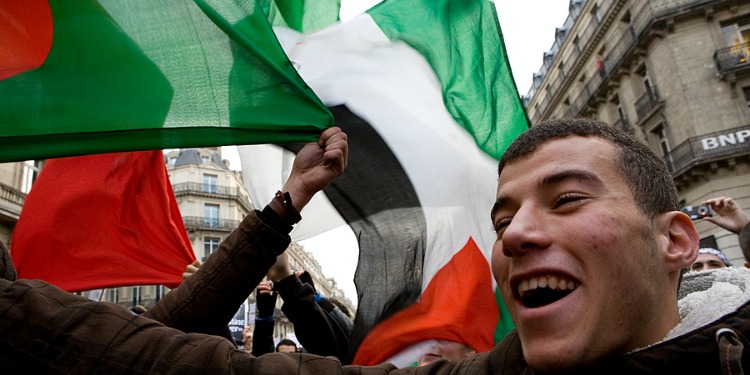 Young man smiling and waving a Hamas flag among a large crowd.