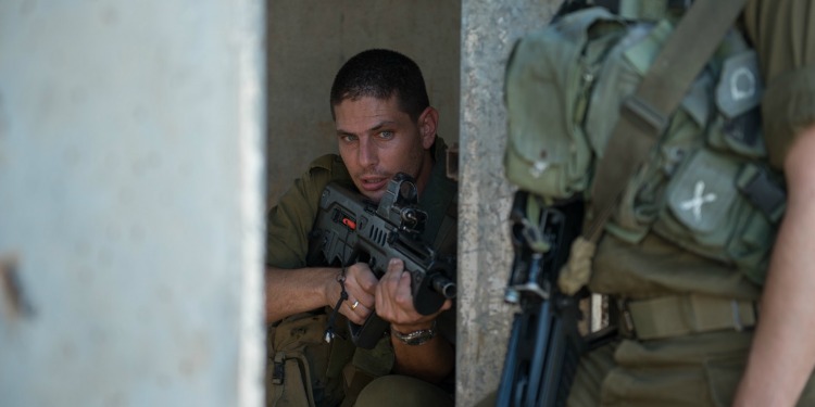 A soldier leaning up against a brick building while holding a gun.