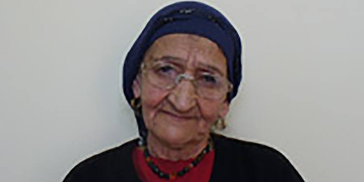 Elderly Jewish woman in a blue headscarf and glasses soft smiling against a white background.